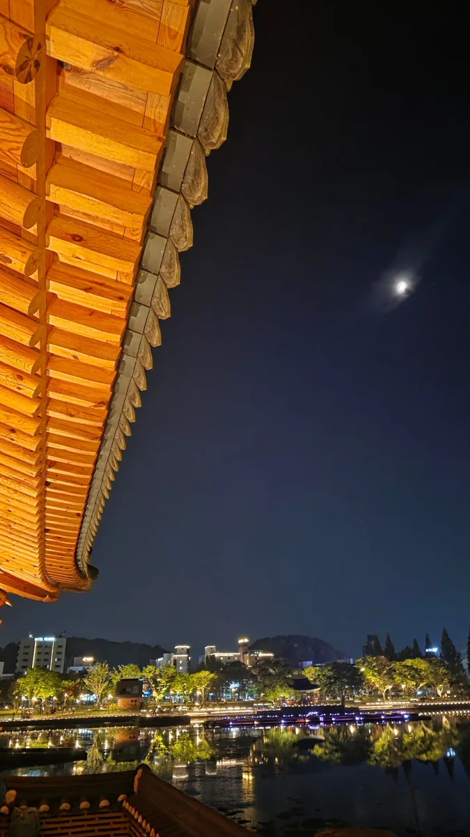 조명을 받아 빛나는 한옥 처마 서까래와 기와, 그 너머 밤하늘의 달